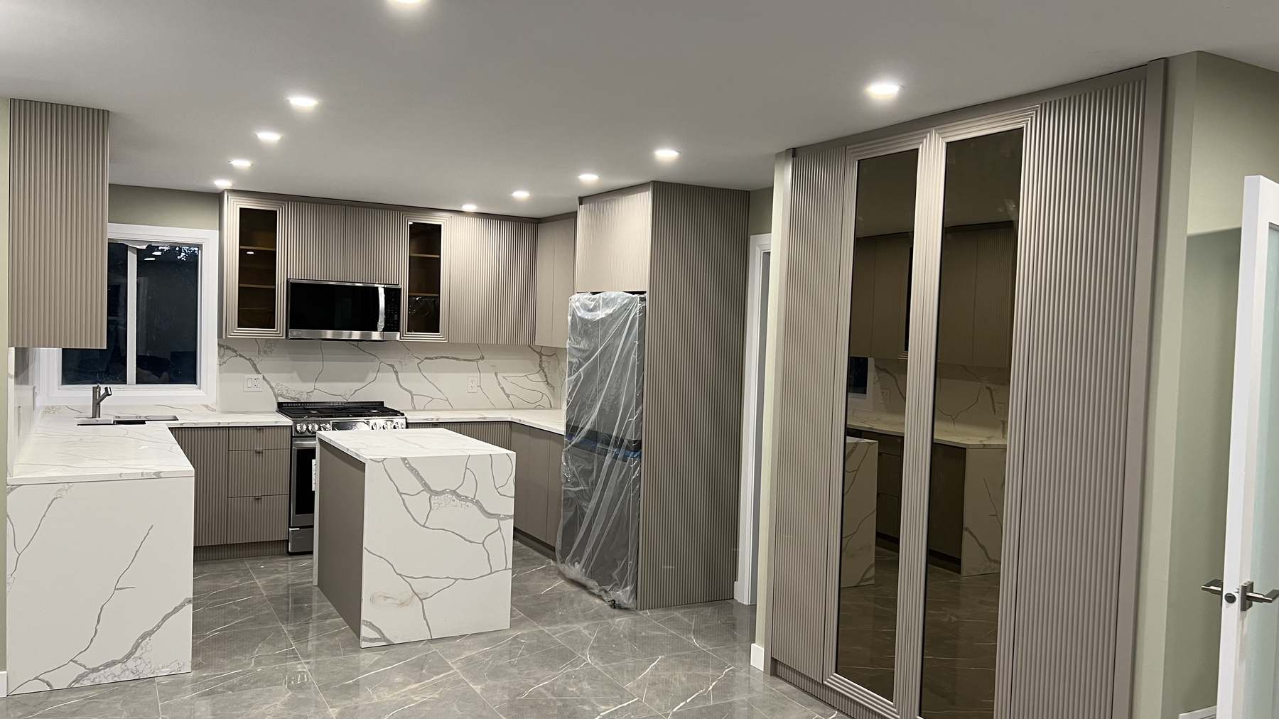 The near-complete kitchen with integrated LED lighting live, glass-front upper cabinets and appliances still wrapped in protection